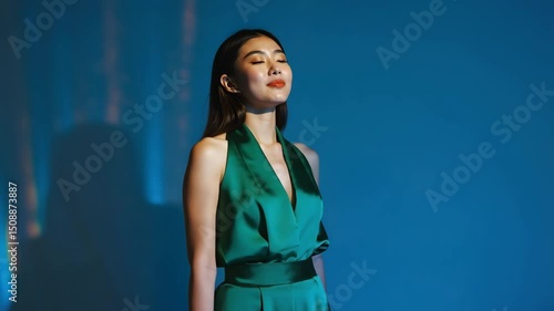 elegant young woman in green satin dress gazing to the left under dramatic lighting on plain blue background, concept of fashion, luxury, evening wear
