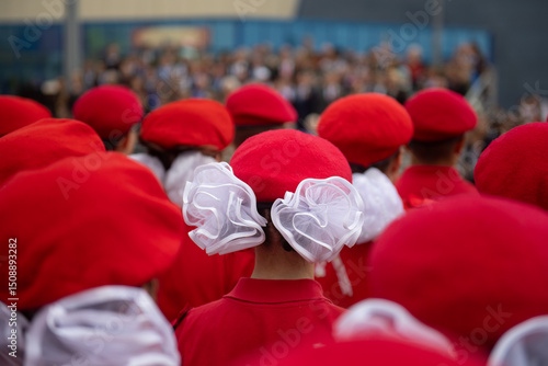 A girl with a white bow in a red beret stands in formation during a parade. The focus is on her back, showcasing the vibrant red uniform and the festive atmosphere of the event.