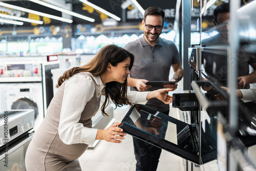 Smiling pregnant woman opening oven door and pointing inside while shopping for new kitchen appliances with cheerful salesman holding digital tablet in electronics store