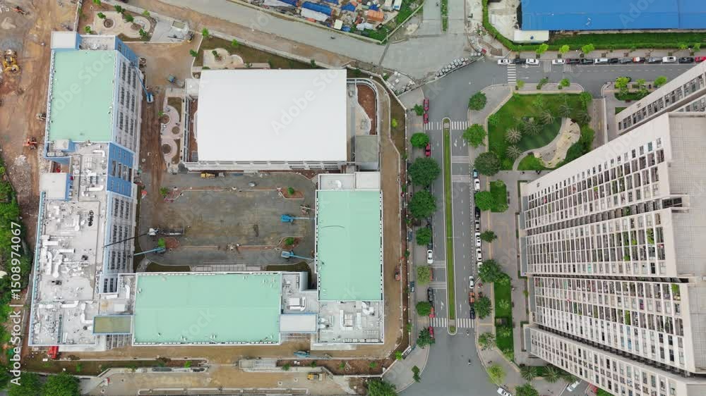 Top-down aerial of a rectangular construction footprint and adjacent crane base, flanked by a white-roofed complex and high-rise tower edge Top-down aerial of a rectangular construction footprint and adjacent crane base, flanked by a white-roofed complex and high-rise tower edge