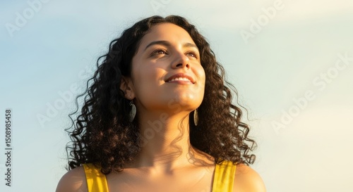 Close-up portrait of a woman looking up with joy against a clear sky background with sun glare, wearing a yellow top, and natural lighting.