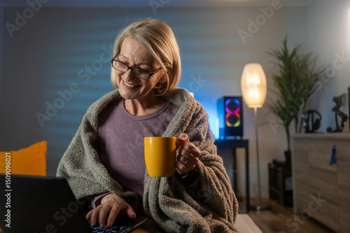 A mature smiling woman wrapped in a cozy blanket drinks tea at home in the living room during the evening, with a laptop nearby, enjoying a peaceful moment.