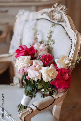 Bridal bouquet with white, pink, and red roses placed on vintage upholstered chair with pearl necklace, lace garter, and wedding veil. Studio still life photography. Wedding preparation and bridal acc