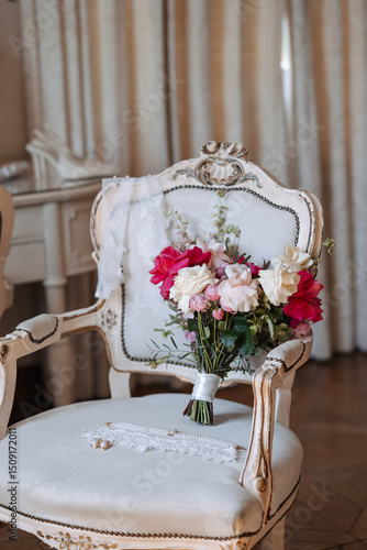 Bridal bouquet with white, pink, and red roses placed on vintage upholstered chair with pearl necklace, lace garter, and wedding veil. Studio still life photography. Wedding preparation and bridal acc