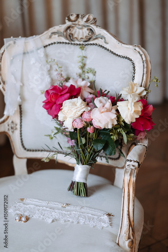 Bridal bouquet with white, pink, and red roses placed on vintage upholstered chair with pearl necklace, lace garter, and wedding veil. Studio still life photography. Wedding preparation and bridal acc