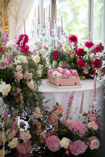 Pink rectangular cake decorated with white and pink ranunculus flowers placed on round white table surrounded by floral arrangements of roses and greenery. Indoor wedding celebration setup. Wedding de