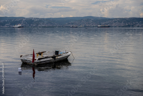 Wallpaper Mural Solitary Fishing Boat Floating on Calm Waters with Cityscape and Cargo Ships in the Distance in Başiskele, Kocaeli, Türkiye Torontodigital.ca