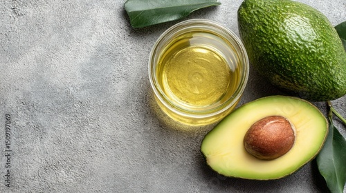 Sliced avocado reveals its creamy interior next to a small bowl of golden avocado oil on a gray textured background with green leaves accentuating the scene