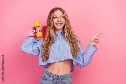 Happy young woman holding a skateboard and pointing to the side wearing stylish blue shirt and glasses against a pink wall photo