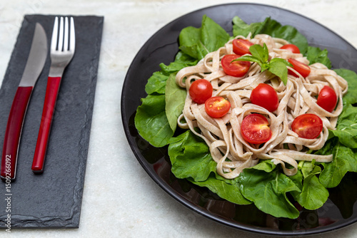 Wholemeal pasta with green leaves of arugula and Cherry tomato on black plate and white marble background