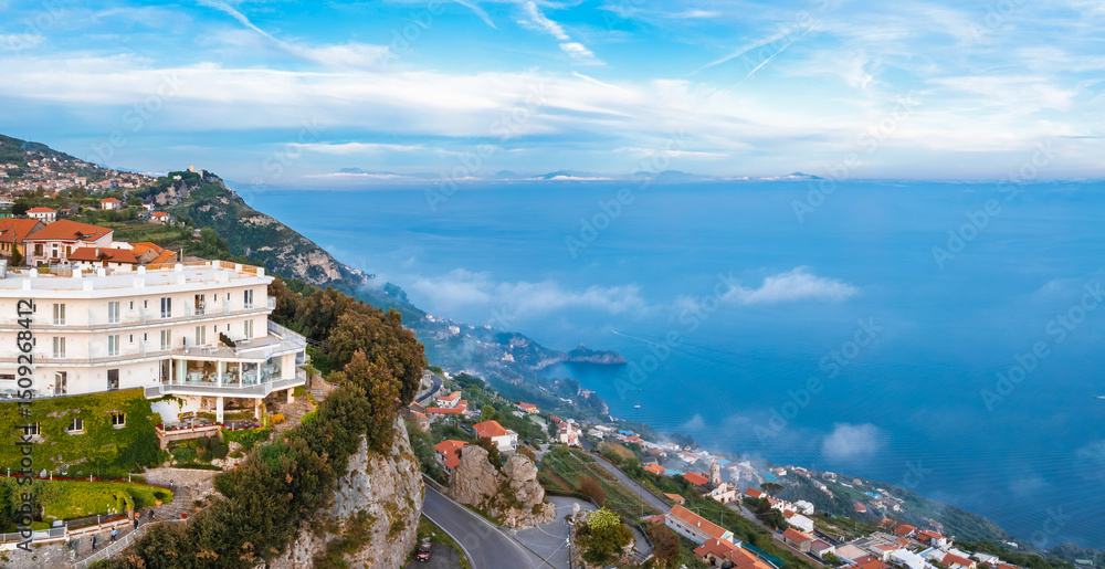 Naklejka premium Aerial view of Amalfi, Italy, featuring a white hotel on a cliff, terraces with greenery, coastal road, red roofed buildings, and the Tyrrhenian Sea.