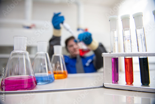 Colorful liquids fill blurred laboratory flasks in the foreground, while a female lab technician in a factory uniform pours red liquid from a pipette into a flask, slightly out of focus, capturing the