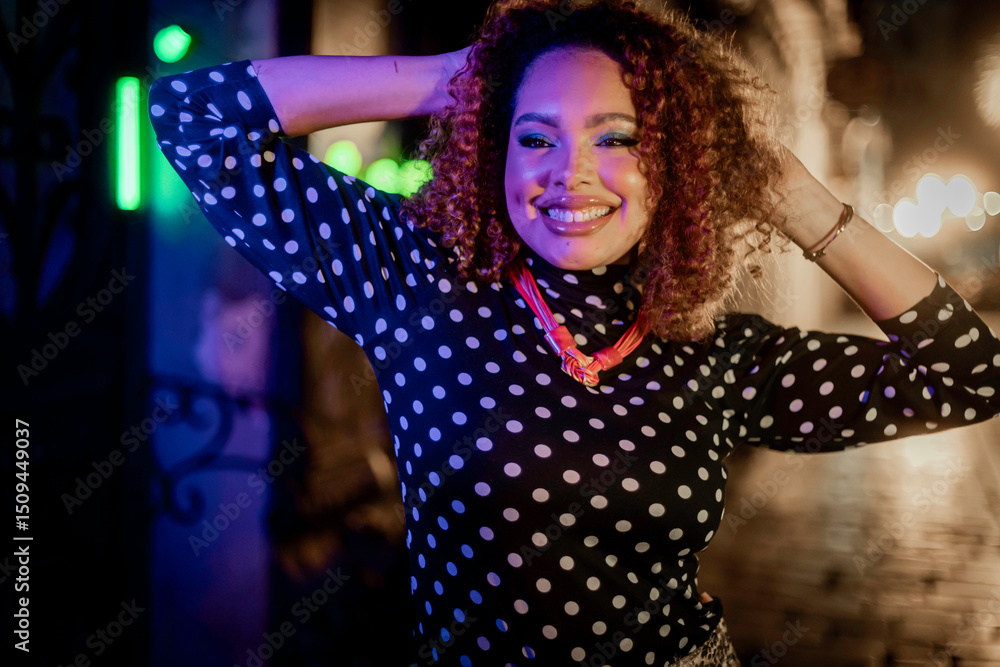Fototapeta premium Black woman with curly hair smiling confidently in a neon-lit street scene inspired by the 1980s. Her striking look with a polka-dot blouse and sparkly skirt conveys style, joy, and bold attitude.