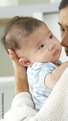 Asian loving young mother gently embraces her three-month-old baby in a cozy room, radiating warmth and affection. Their tender connection fills the space with a sense of comfort, security, and love.