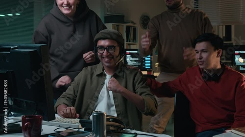 Medium shot of obnoxious multiethnic accomplices standing around Indian guy typing on computer, attentively looking at screen, then cheering at scamming victim of life savings in identity theft