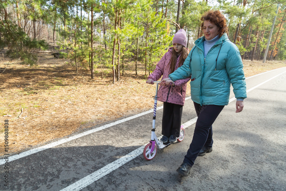 Little girl riding scooter while walking with her grandmother in the forest on a sunny day Little girl riding scooter while walking with her grandmother in the forest on a sunny day