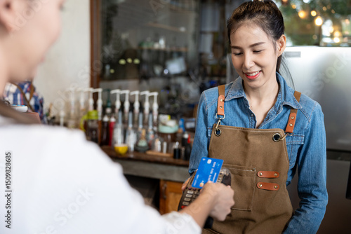 Asian female barista in denim shirt and brown apron smiling while holding card reader as customer pays with credit card. Friendly cashless payment interaction inside modern coffee shop.