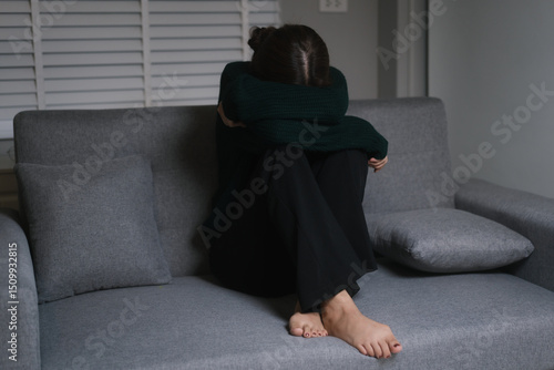 Canvas Print A young woman, who appears to be of Asian descent, sits curled on the floor with a melancholic expression, leaning against gray sofa, conveying feelings of loneliness and sadness in dimly lit room