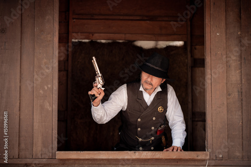Fotografie Portrait elder cowboy standing at a wooden window frame, aiming a silver revolver upward in a dramatic on the weapon suggest respond to danger, possibly during a shootout or high-stakes standoff