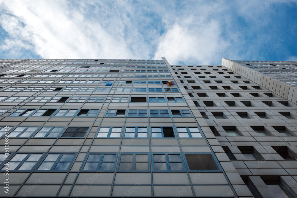Upward view of a high-rise residential building with a repetitive grid of square windows and light-colored panels. Upward view of a high-rise residential building with a repetitive grid of square windows and light-colored panels.