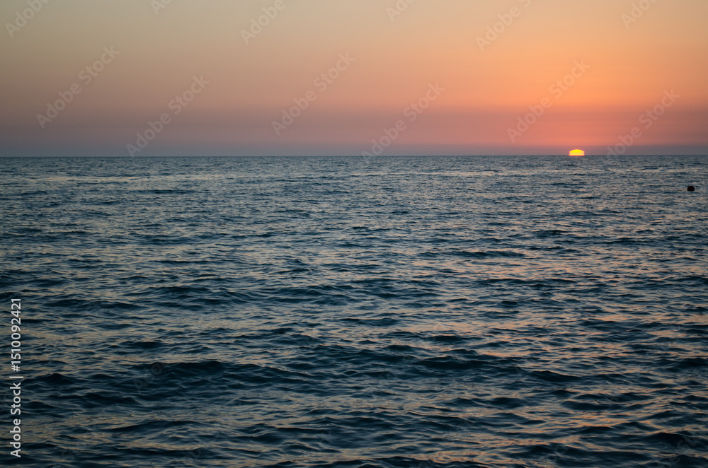 Obraz premium Calm ocean scene under warm, gradient sunset sky. Sun partially dips below horizon, casting orange glow. Peaceful atmosphere with gentle waves and expansive view