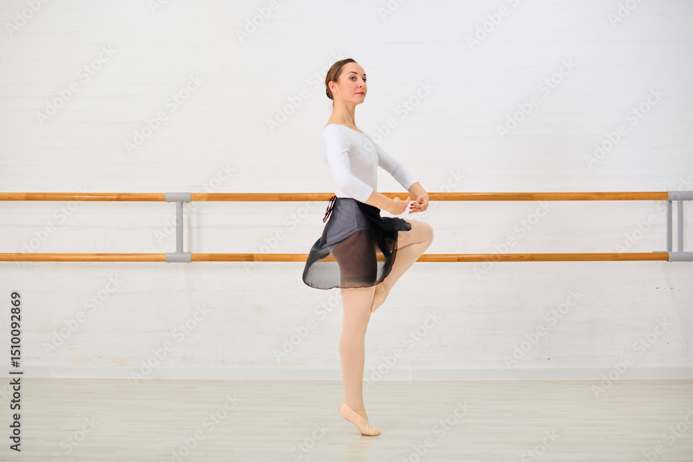 Naklejka premium Young female ballerina with brown hair in white top, black skirt, and pink tights, practices moves in bright studio. Wooden barres line neutral-toned wall