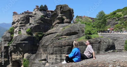 Attractive young couple sits on a cliff edge with a Meteora monastery in the background
