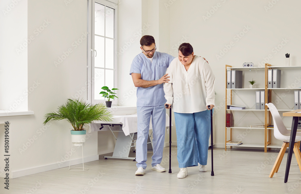 Fototapeta premium A young cheerful friendly nurse helping fat young patient to walk with her crutches in medical clinic. Male physiotherapist or health care worker helping woman in rehab.