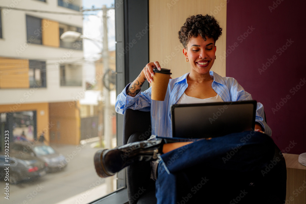 Obraz premium Smiling young creative woman with tattoos is comfortably working remotely, enjoying a coffee while using a digital tablet in a modern office space with large windows and city views