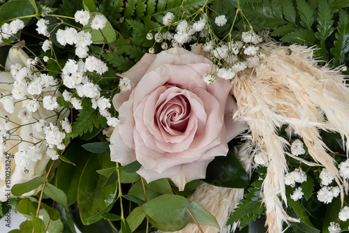 Floral arrangement featuring a soft, dusty pink rose at the center surrounded by delicate white baby's breath flowers, creamy pampas grass, and rich greenery part of a wedding bouquet or a centerpiece