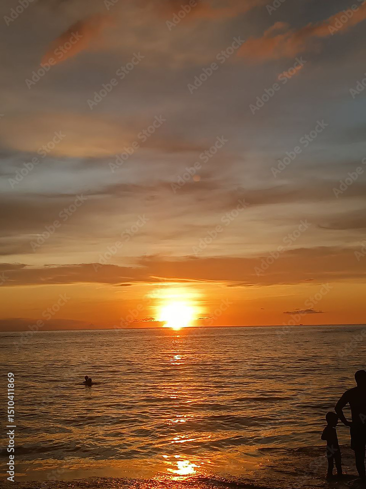 Fototapeta premium Seascape photography. Thick horizontal clouds. Tropical sky in orange colors. Panorama of the southern sea. Sun path in the water. Sunset ocean landscape for wallpaper. Human silhouettes on the shore.
