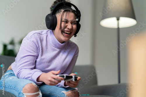 A happy young woman with headphones plays a video game on a console, holding a controller and laughing loudly while sitting on a comfortable sofa at home.