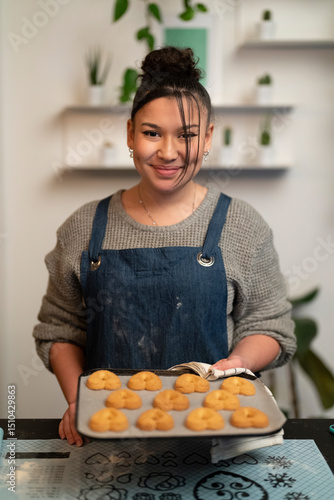 A young woman wearing a denim apron holds a baking tray filled with freshly baked heart-shaped cookies. She stands in a kitchen and smiles proudly at the camera, showcasing her homemade treats.