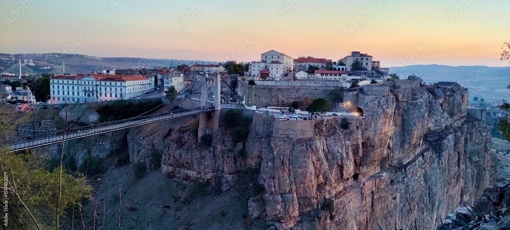 Fototapeta premium the iconic Sidi M'Cid Bridge and the city's dramatic cliffs. The setting sun bathes the rugged landscape and historic buildings in warm light, showcasing the 