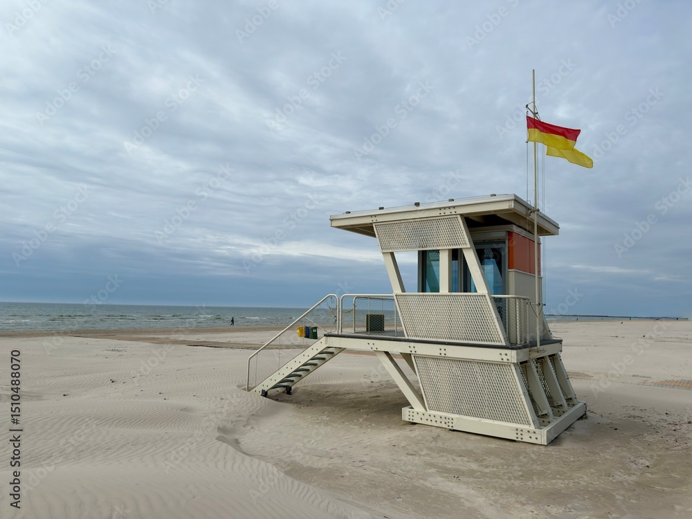 Obraz premium miami beach lifeguard tower, Modern lifeguard tower on a wide sandy beach by the sea under a cloudy sky. A red and yellow flag is flying, signaling beach safety conditions in a peaceful coastal scene