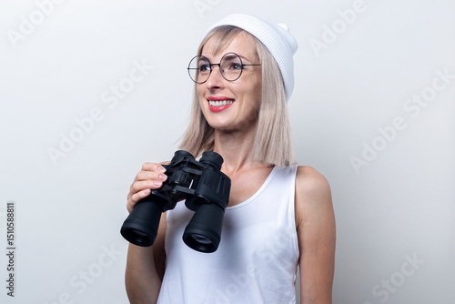 Cheerful young woman holding binoculars looks to the side on a light background