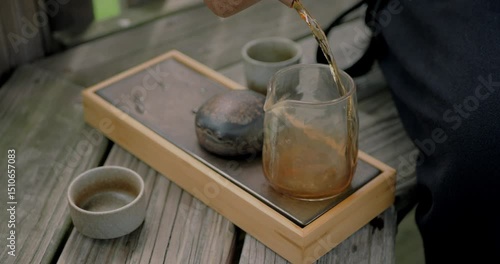 Close-up of traditional Chinese tea being poured into a glass cha hai on a bamboo tea tray with ceramic cups. Peaceful outdoor setting reflecting authentic tea ceremony and mindfulness.