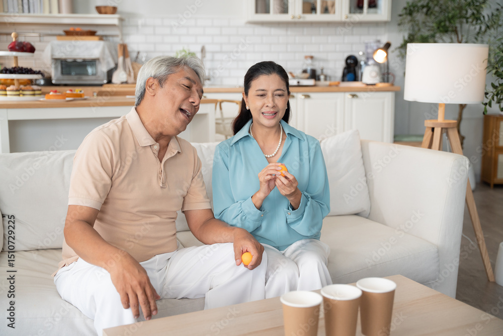 Asian senior couple playing toss ping pong ball to the cup in the living room at home on weekend after retirement. Healthy insurance concept Asian senior couple playing toss ping pong ball to the cup in the living room at home on weekend after retirement. Healthy insurance concept