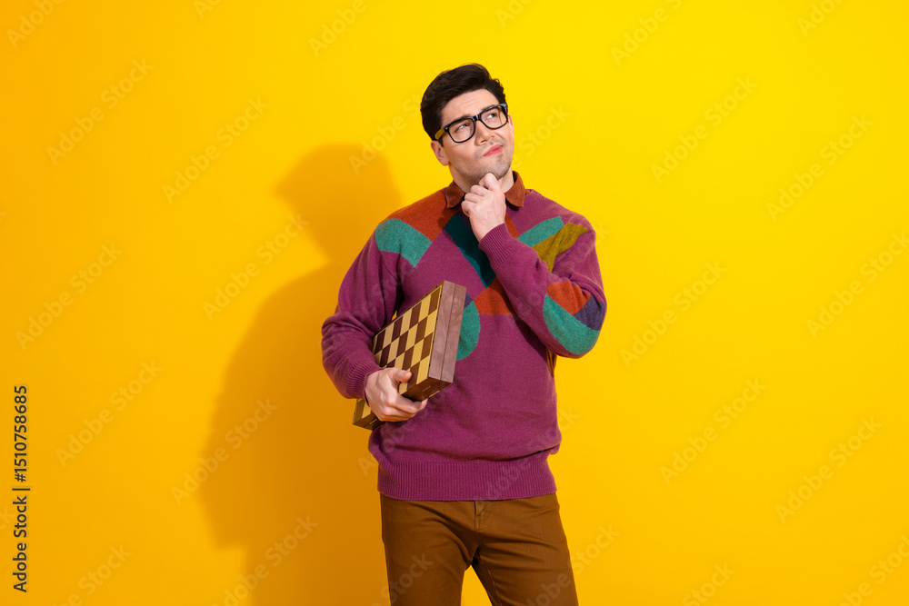 Fototapeta premium Young man in colorful sweater holding checkered game board, pondering decision against vibrant yellow background
