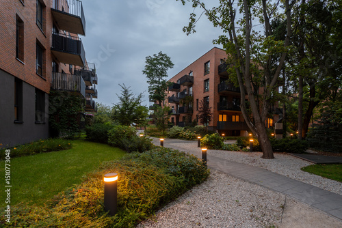 Illuminated pathway in landscaped courtyard of modern residential complex at dusk. Bollard lights create warm ambiance among lawns, shrubs, trees, near apartment buildings. A peaceful community space.