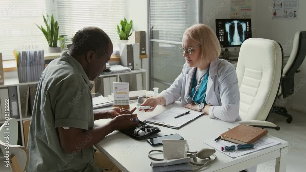 African American man pricking his finger with pen while female clinician with glucometer monitoring his blood sugar level and showing him data on device screen during medical consultation African American man pricking his finger with pen while female clinician with glucometer monitoring his blood sugar level and showing him data on device screen during medical consultation
