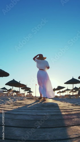 Fashionable woman in a white dress enjoys walking beach wooden pathway at sunrise, vertical video