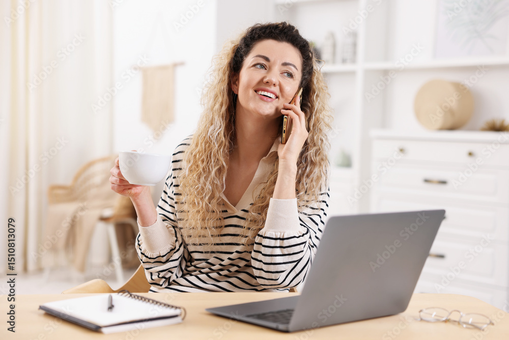 Smiling woman talking by smartphone at desk with laptop in home office Smiling woman talking by smartphone at desk with laptop in home office