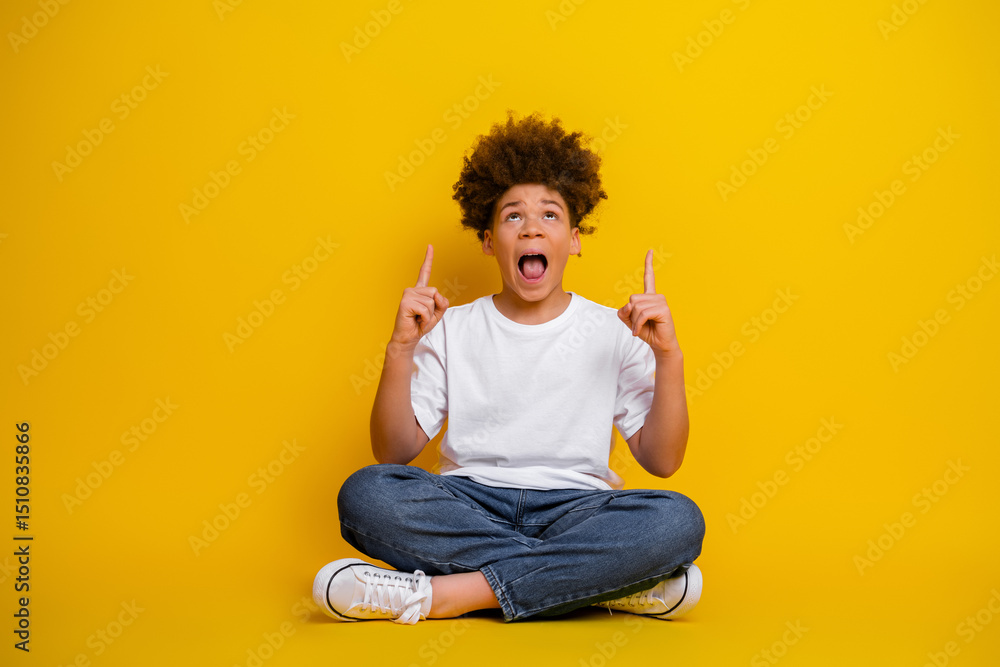 © deagreez - Excited young boy pointing upward while sitting cross-legged against a bright yellow background in casual attire