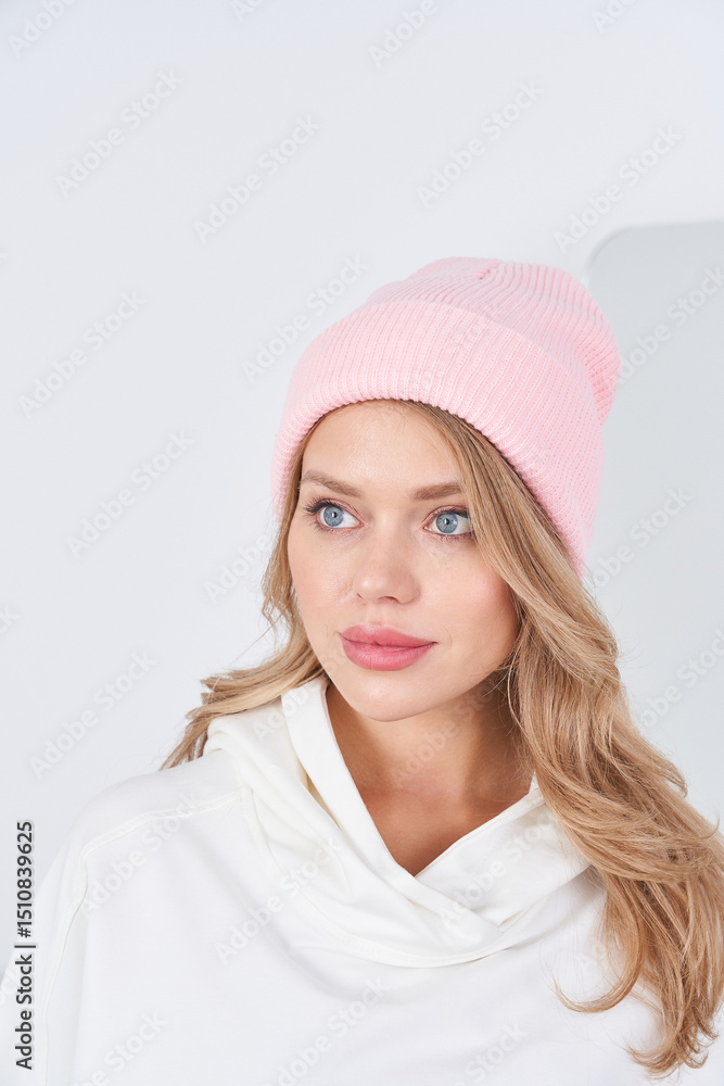 Fototapeta premium Young woman with long hair wearing a pink knit beanie and a white cowl-neck top, looking to the side. Soft lighting enhances the serene and stylish appearance