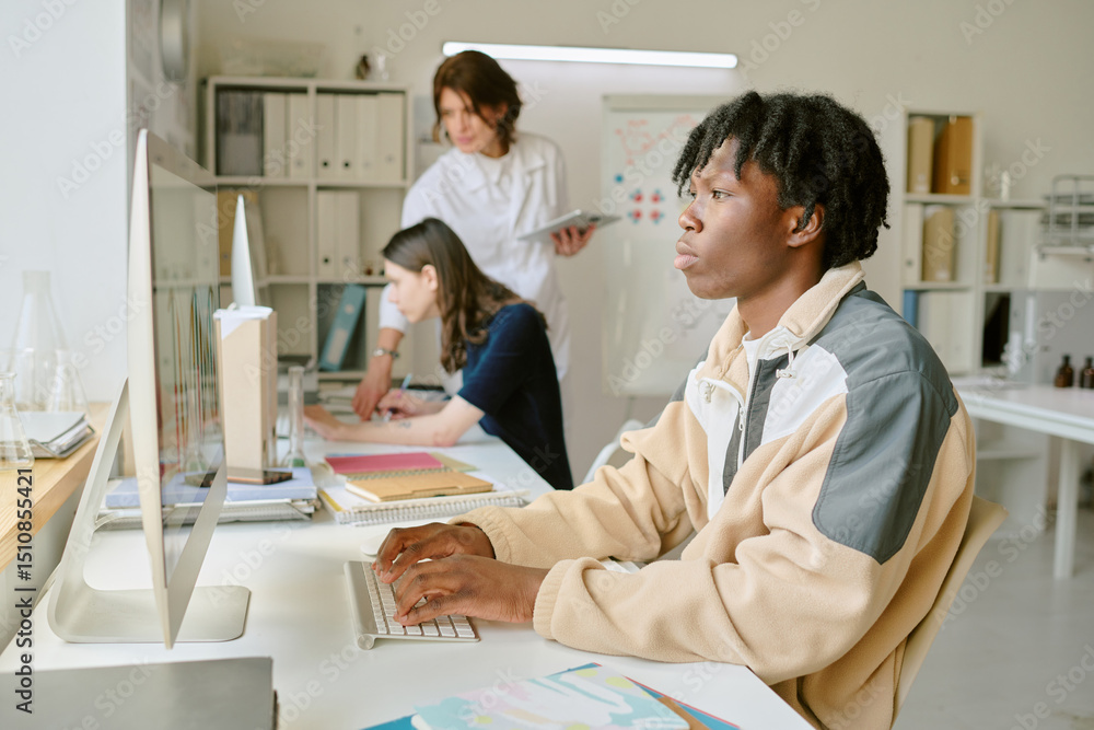 Obraz premium Student working on computer, focused and typing while two colleagues collaborate in background in bright, organized office space featuring various workstations and charts