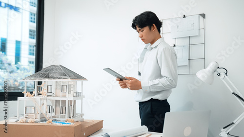 Canvas Print Professional handsome asian project manager using tablet to check house model while inspect house model structure carefully at office with blueprint hanged behind