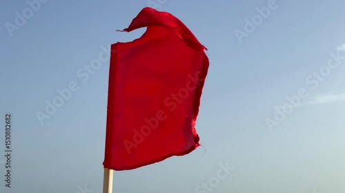 A red flag flutters prominently at the beach, warning swimmers of dangerous water conditions, signaling to beachgoers to avoid entering the sea. The sky is clear and sunny.