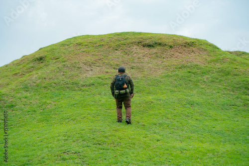 A male adventurer is enjoying the beauty of a small hill covered in green grass. The Asian male traveler is relaxing on the clean and beautiful green hills.