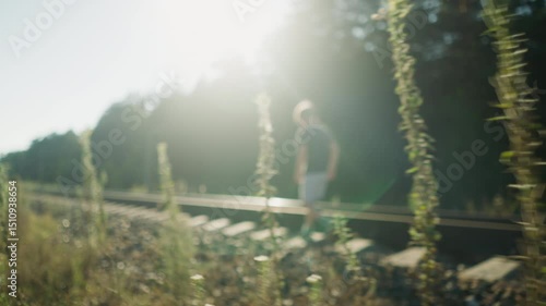 Little boy walking along rail track with bright sunlight casting strong glow through surrounding trees and wild plants, creating dramatic silhouette and peaceful rural atmosphere